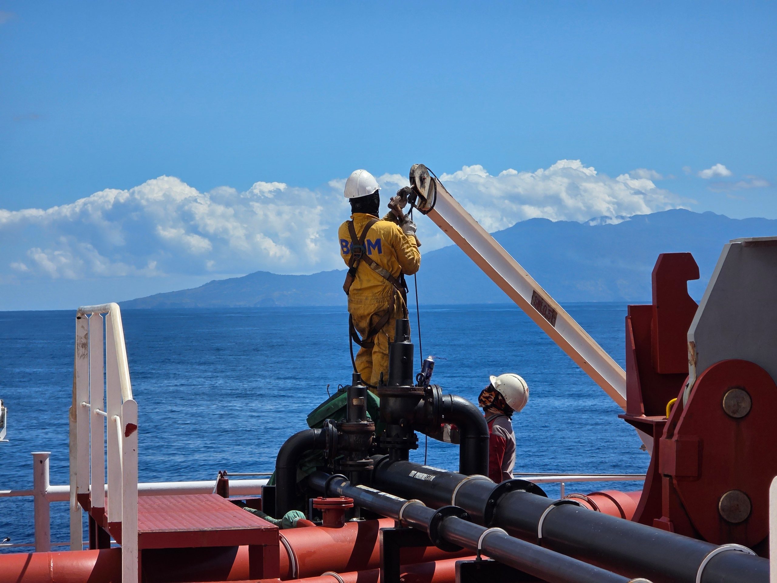 Two marine workers operate equipment on an offshore vessel in Mozambique, scenic ocean and island backdrop.